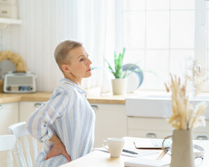 Tired exhausted middle aged business woman massaging her back while sitting in kitchen and working or studying on laptop online from home. Senior people and technologies concept