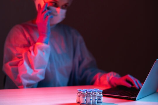 Covid-19 vaccine. Online research. Innovative technology. Female scientist in ppe working on laptop speaking on mobile phone with vial dose bottles in red blue neon light on dark blur background.