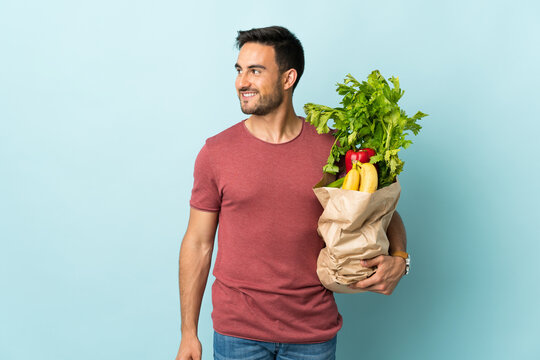 Young caucasian man buying some vegetables isolated on blue background laughing