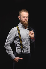 Portrait of Caucasian attractive bearded man in stylish suit and shirt with suspenders looks into the camera while holding jacket on shoulder. On a black background.