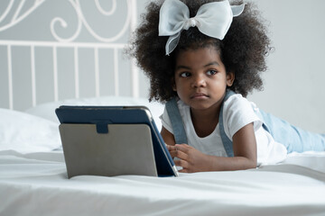 Little cute African kid lying down on bed absent minded and looking other way while using tablet computer to learn online class at home