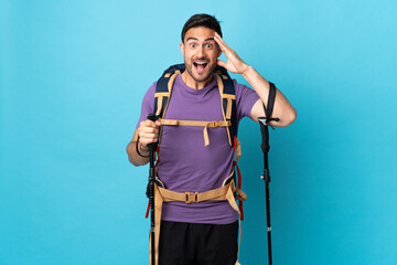 Young caucasian man with backpack and trekking poles isolated on blue background with surprise expression