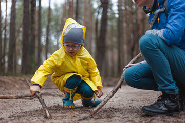 Mom and child walking along the forest road after rain in raincoats, together draw on the sand with wooden sticks