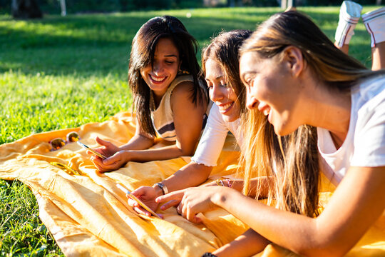 Three young happy women having fun with internet mobile technology using smartphone lying on meadow in city park at sunset or dawn. New habits of girls sharing life moments, photo or contacts