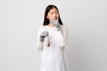 Chinese fishmonger wearing an apron and holding a raw fish over isolated white background having doubts and with confuse face expression