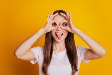 Photo of funny crazy lady two okey signs cover eyes protrude tongue wear white t-shirt posing on yellow background