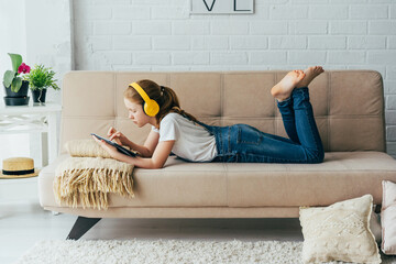 Red-haired girl child lying on the couch with an electronic tablet listens to music in yellow headphones. Remote learning, teenager using wireless devices doing homework or watching social networks.