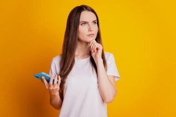 Photo of minded lady hold phone think look empty space wear white t-shirt posing on yellow background