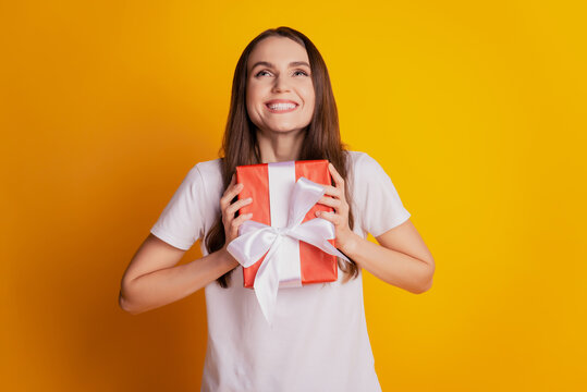 Photo of nice glad emotional lady hold giftbox look up empty space wear white t-shirt posing on yellow background