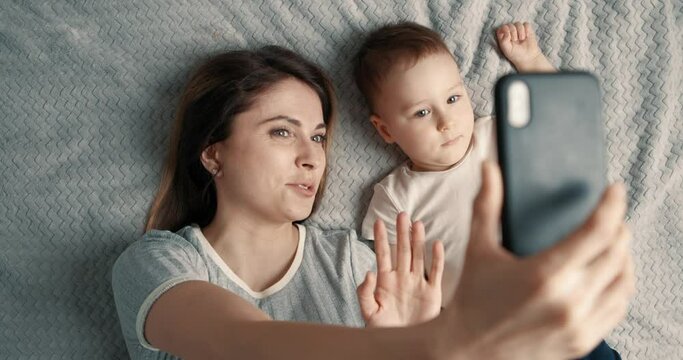Smiling woman and baby boy making a selfie or video call in a bed and waving their hands. Concept of technology, new generation, family, connection, parenthood. Top view, slow motion.