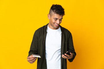 Young brazilian man isolated on yellow background buying with the mobile with a credit card