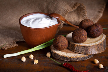 Close up with fried falafel on wooden circles and white yogurt sauce in clay bowl on brown board with burlap cloth. Healthy roasted vegetarian food. Jewish cuisine. Green onion and chickpea