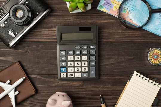 Top view photo of calculator in the center piggy bank plane model camera map magnifier compass passport cover pen plant and notebook on isolated wooden table background