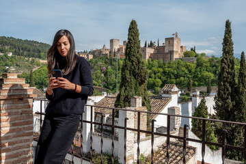 Hispanic female tourist messaging on mobile phone while standing on terrace with views to Alhambra fortress in Granada, Spain
