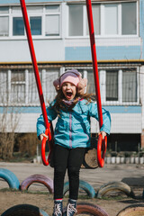 A funny, smiling girl, a preschool child rides, sitting on a metal red swing, on the playground. Happy childhood.