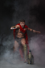 Fototapeta na wymiar Young athletic sexy man in a costume of a Roman warrior in a red cloak in move on a black background in the smoke.