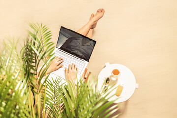 Woman freelancer with laptop sitting under palm tree branches. Sunscreen, sunglasses, orange juice on the table of sandy beach . Summer vacation. Top view