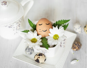 An Easter egg with a painted peaceful face, lying in a white porcelain cup in green leaves and white flowers on a white background. The concept of the Easter holiday. Copy the space for the text.