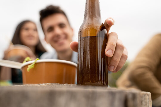 Guy pick up beer from the table outdoors. Closeup on the bottle with no label. Young man eating take away food outdoors and having fun drinking beers together. blurred people faces on the background..