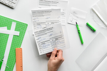 Obraz premium Fake covid vaccination record card. Covid scams. Forged health certificate with false vaccination record against covid-19. Blank card, pen, paper knife,laminator. Top view, flat lay.