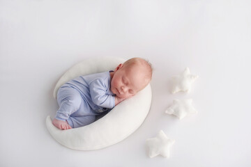 A newborn baby boy in a knitted suit sleeps on the white moon next to the white stars on knitted blankets