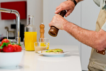 horizontal view of unrecognizable man preparing and avocado toast with pepper sprinkled with a shaker. Spanish breakfast preparation with fresh vegetables. Food and cooking concept.