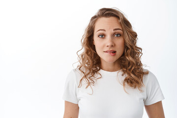 Fototapeta premium Nervous blond girl biting lip and looking scared, making silly mistake, looking worried at camera, standing against white background