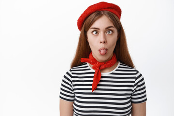 Close up of funny girl showing silly face and tongue, squint eyes and fool aruond, stands in french outfit, white background