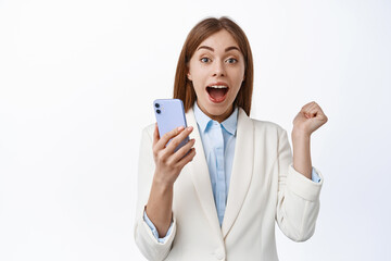 Fototapeta premium She is winner. Lucky corporate woman scream from joy, winning on mobile phone, triumphing from good news in internet, holding cellphone and jumping from happiness, white background