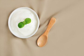 Top view of, Yogurt natural flavor in white ceramic bowl with peppermint leaf and wooden spoon placed on light brown cloth with copy space, Milk curd organic homemade for health care and healthy food.