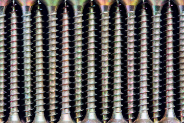 Golden brass steel wood tapping screws galvanized with rainbow tint lined ornamental macro background pattern, lying in a row over black surface mirror-like relative to each other, top view closeup.