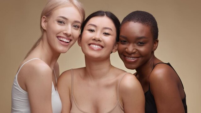 Studio portrait of three happy multiethnic ladies laughing to camera, enjoying friendship, hugging over beige background