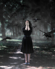 Fine art outdoor portrait of beautiful caucasian young woman in black dress. Girl standing alone on a forest path, covering her face with her hands, surrounded by a flock of flying crows