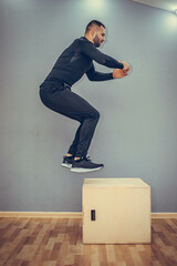 Man jumping onto the box on gray wall background. Fit athletic man jumping up to plyometric wood box, home workout exercise concept.