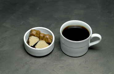 black coffee in a white cup and chocolates in a small white saucer on a gray background
