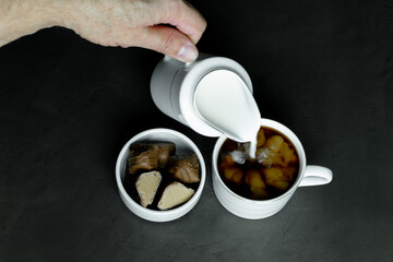 a female hand pouring milk into black coffee in a white cup, nearby are chocolate candies in a small white saucer on a black background