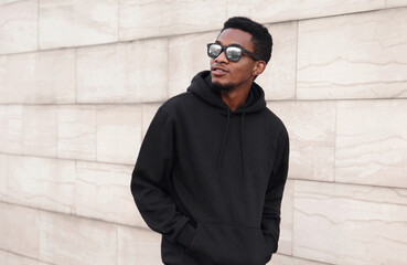 Portrait of stylish young african man walking and looking away wearing a black hoodie, sunglasses on a city street over gray wall background