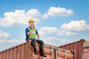 Foreman worker in hardhat and safety vest sitting on container box talks on two-way radio for...
