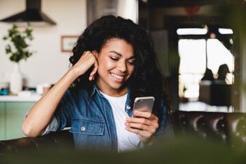Cheerful african teenage girl using smart phone sitting on the sofa.Smiling african american woman using smartphone at home, messaging or browsing social networks while relaxing on couch