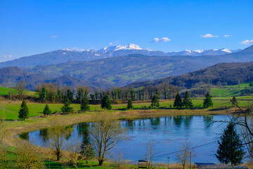 Beautiful spring landscape with mountain lake and mountains covered with snow across blue sky. Fir trees, and trees reflected in water.