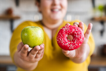 Chubby fat woman holding green apple and sweet donut selective focus. Choice between healthy and...