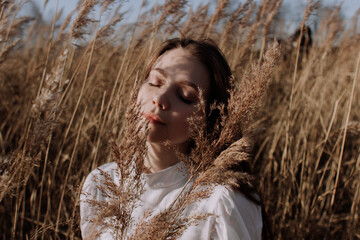 Young woman with closed eyes in white blouse sitting in field of pampas grass in front of sky and sun. Style and street fashion. Girl in casual outfit looking at camera. Golden hour. Shadows on face