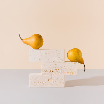 Golden yellow pears balancing on travertine marble blocks structure against gray and beige background. Creative gold summer fruit arrangement. Minimal retro styled surreal food art concept.