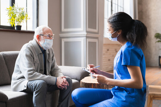 Young woman african doctor give a consultation to elderly old senior grandfather man during home visit in medical mask against Covid 19. Woman nurse visit patient at home consult about insurance.