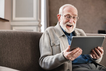 Laughing senior old grandfather using digital tablet on the sofa . Modern elderly pensioner reading e-book indoors with positive emotions