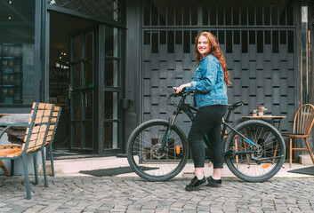 Fashion portrait of red curled long hair caucasian teen girl walking with bicycle on the city...
