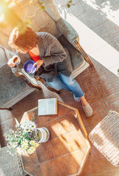 Top view of a female dressed in cozy home clothes sitting in a comfortable armchair and drinking morning coffee in house sunroom living room while she reading a book. Stay home concept image.