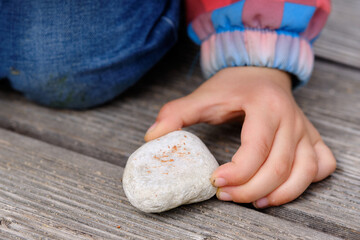 Close-up of the hand of a 4 year old caucasian child holding a white stone in her hand between her fingers on a wooden ground. Seen in April in Germany.