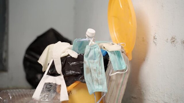 Close up of healthcare worker or doctor throwing single use disposable medical face mask and gloves into filled trash can or garbage bin at hospital during coronavirus covid-19 pandemic.