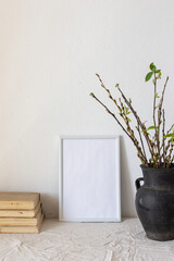 Home decor mock-up, blank picture frame near white painted concrete wall , black earthenware jug with pussy willow branches and blossoming green leaves, old books